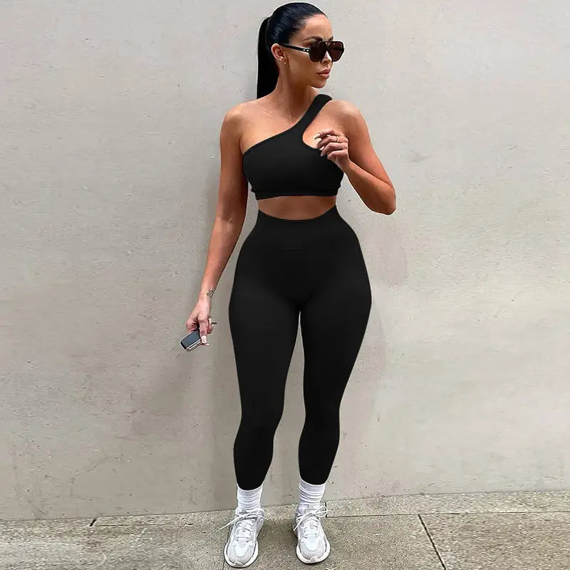 Woman posing in a black one-shoulder crop top and high-waisted leggings with white sneakers.