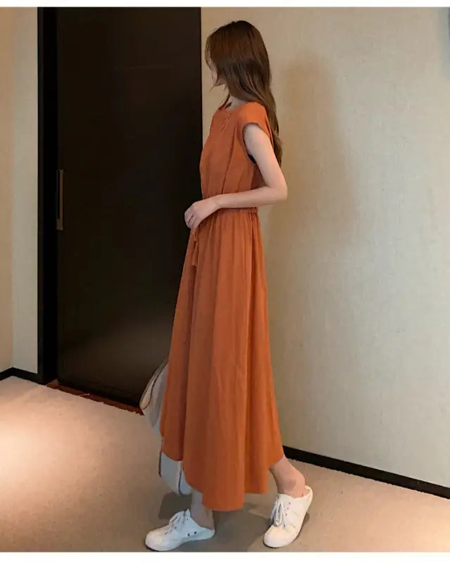 Long orange sleeveless dress with a flowy skirt worn over light-colored pants and white sneakers.