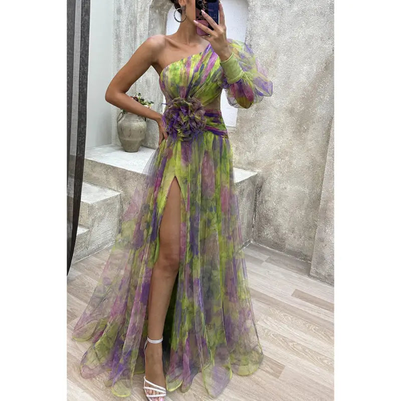 Flowing one-shoulder gown in sheer fabric with green, purple, and pink floral patterns, featuring a high slit and gathered waist detail.