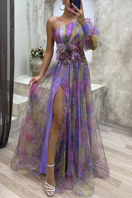 Flowing one-shoulder gown in sheer purple, pink, and gold tones with a high slit and feathered waist detail.