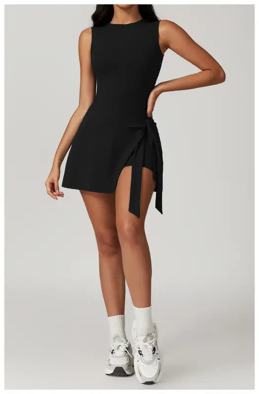 Black sleeveless mini dress with a side slit and tie detail, paired with white socks and chunky sneakers.