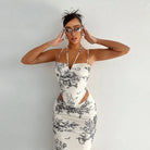 Woman wearing a form-fitting white dress with black floral print and cutout details, posing with hands near her face and stylish sunglasses.