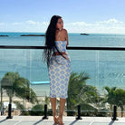 Woman with long dark hair wearing a blue and white floral outfit on a balcony overlooking the ocean.